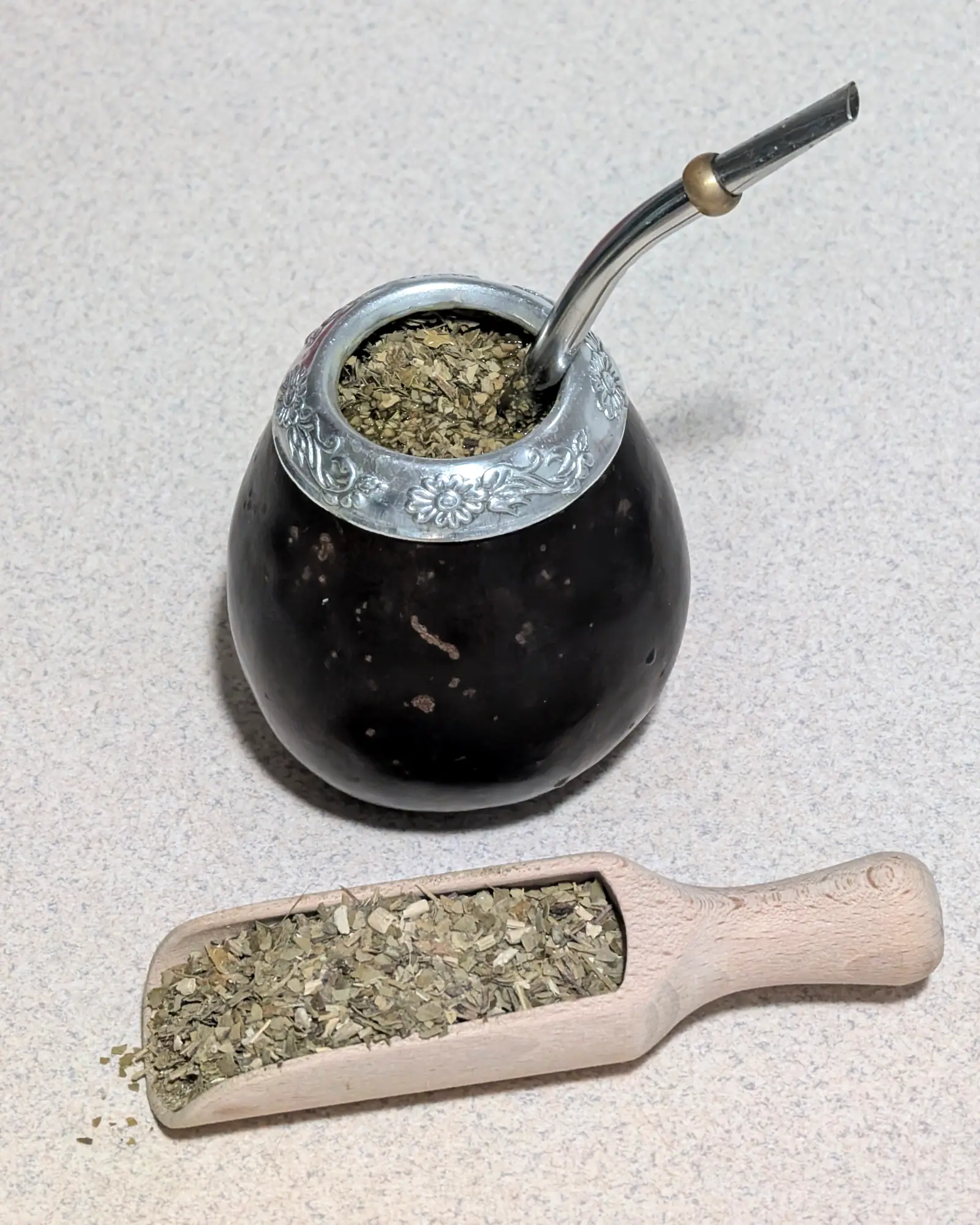 A traditional calabash gourd filled with yerba mate leaf and a metal bombilla straw, beside a small scoop of loose yerba