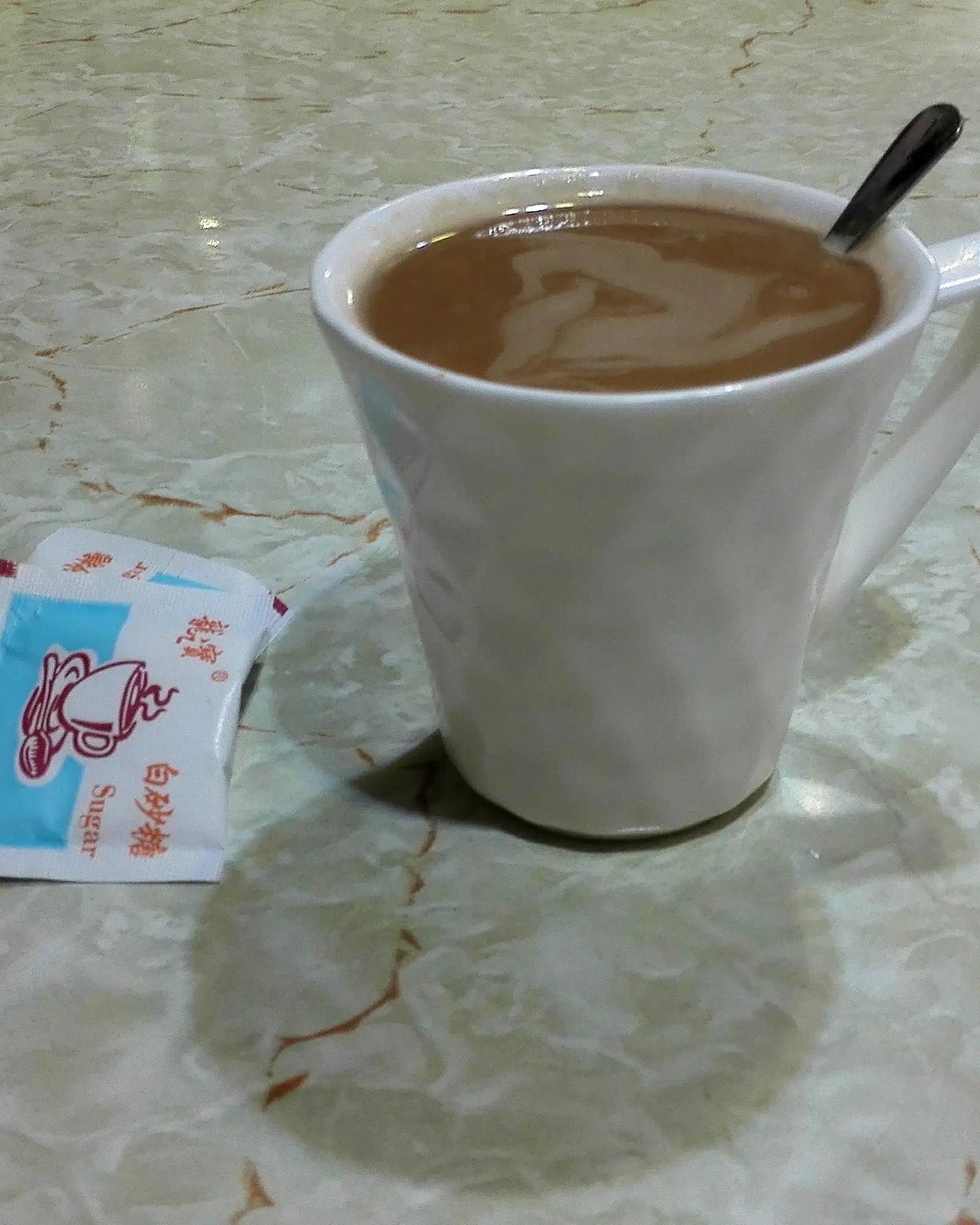 A white mug of milky tea-coffee yuanyang with a spoon resting inside, on a marble table