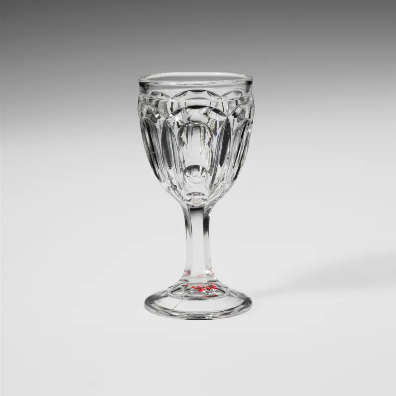 An empty small clear pressed-glass cordial glass with a faceted bowl and short stem, on a plain background