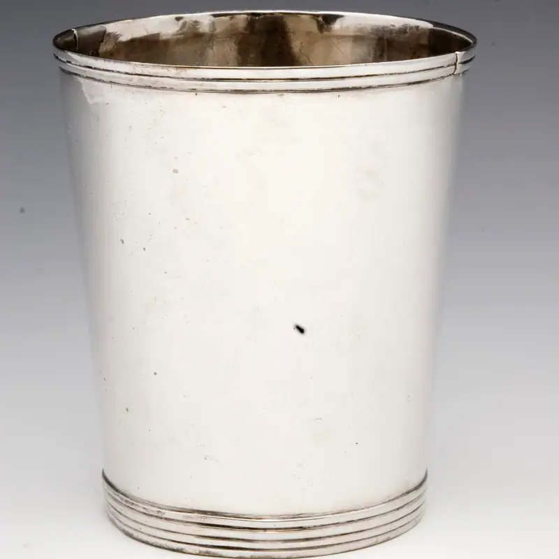 An empty, polished silver julep cup with a tapered shape and ribbed base, on a plain background