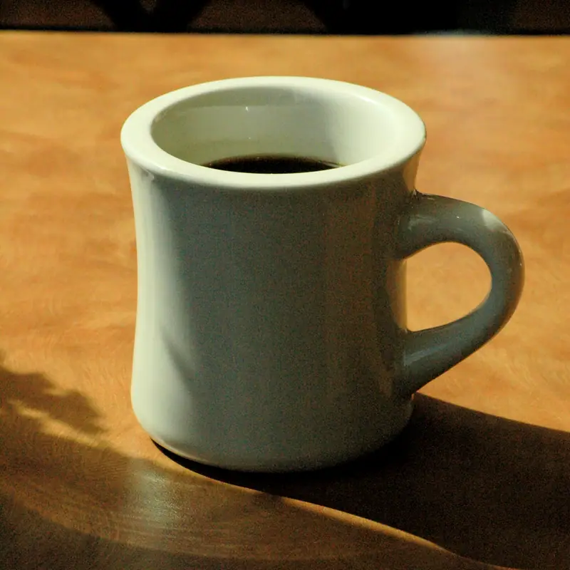A thick white ceramic mug of black coffee with a rounded handle, standing on a wooden table