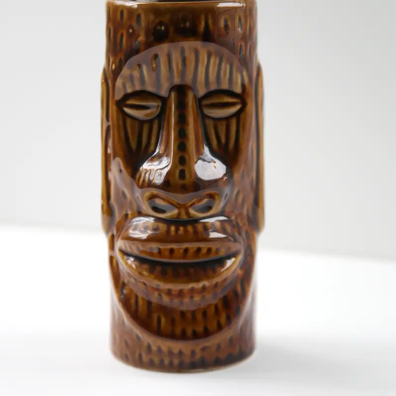 An empty brown ceramic tiki mug carved with a stylised face, on a plain background