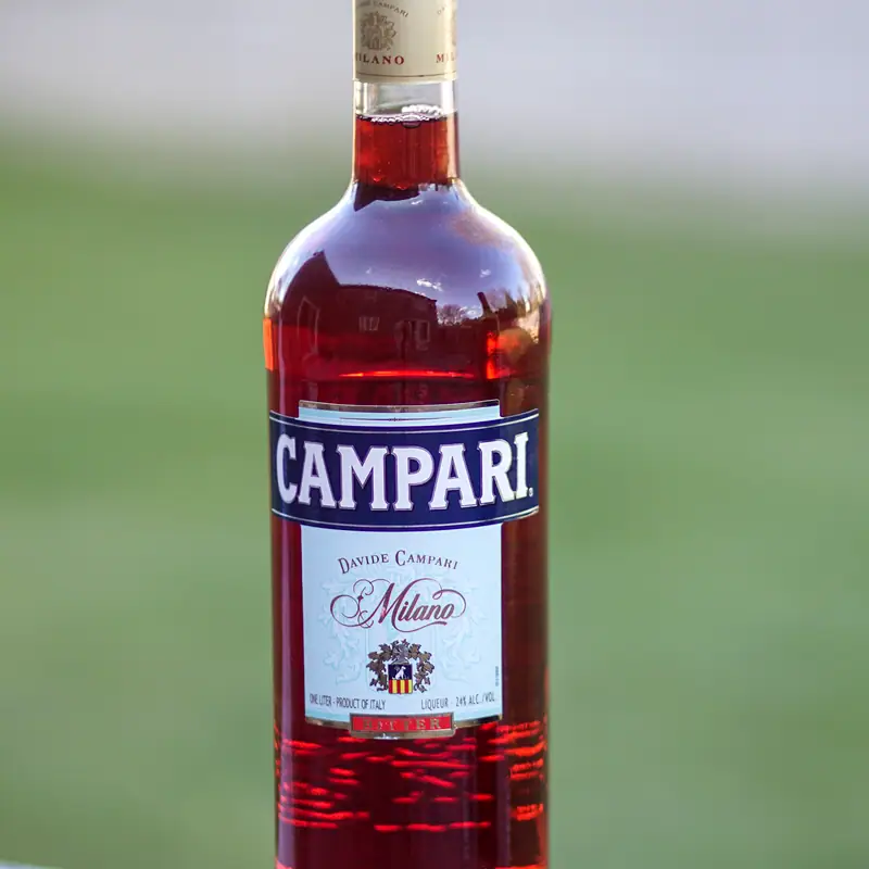 A bottle of Campari, deep red behind clear glass, with its white and blue label, on a green background