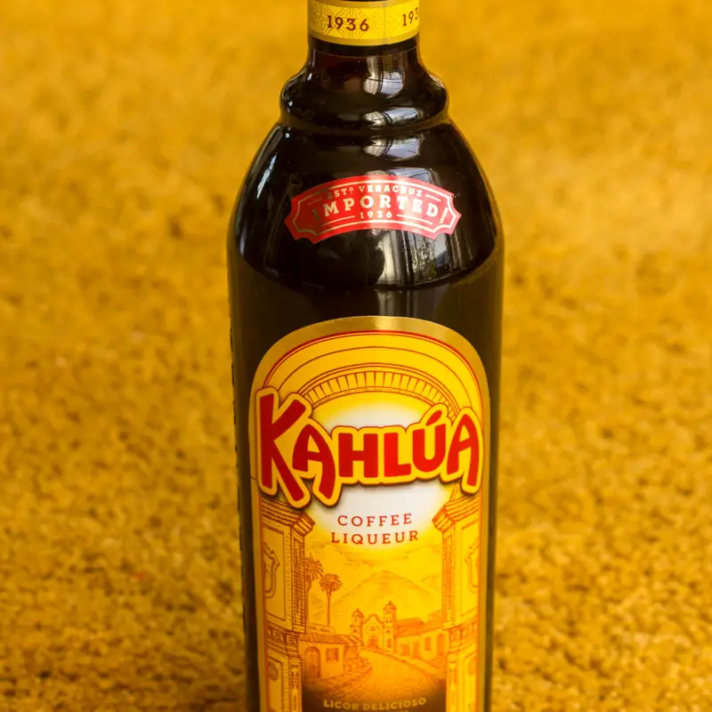 A bottle of Kahlúa coffee liqueur, dark glass with a yellow and red label, against a golden background