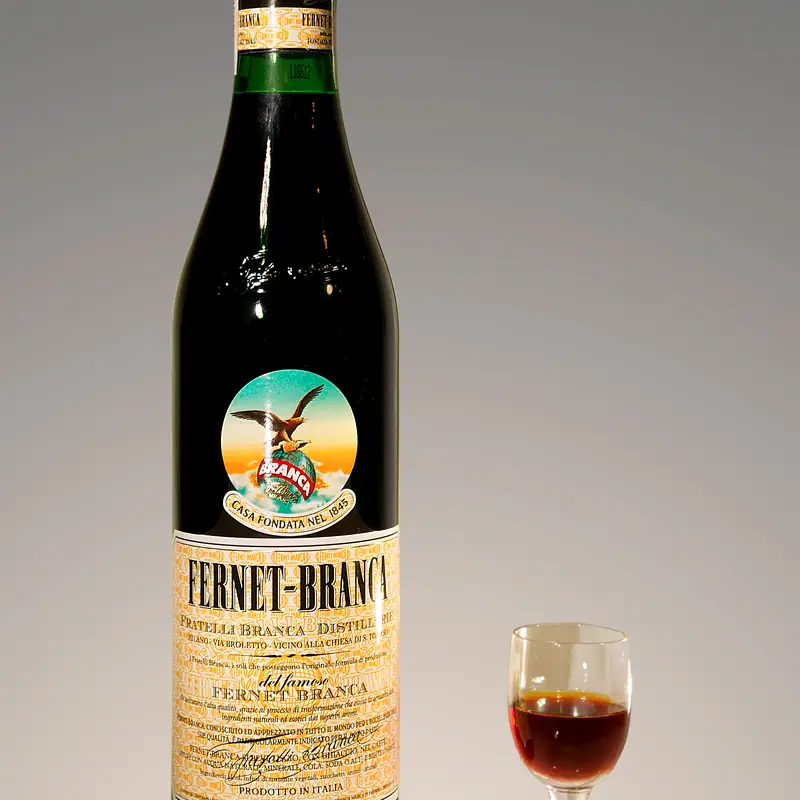 A bottle of Fernet-Branca with its cream label and eagle emblem, beside a small glass poured from it, against a grey studio background.