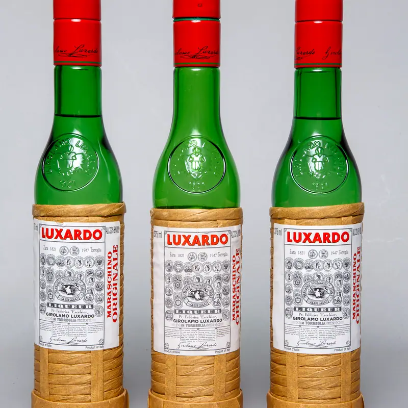 Three bottles of Luxardo Maraschino liqueur, green glass wrapped in straw with a red cap, side by side against a plain background.