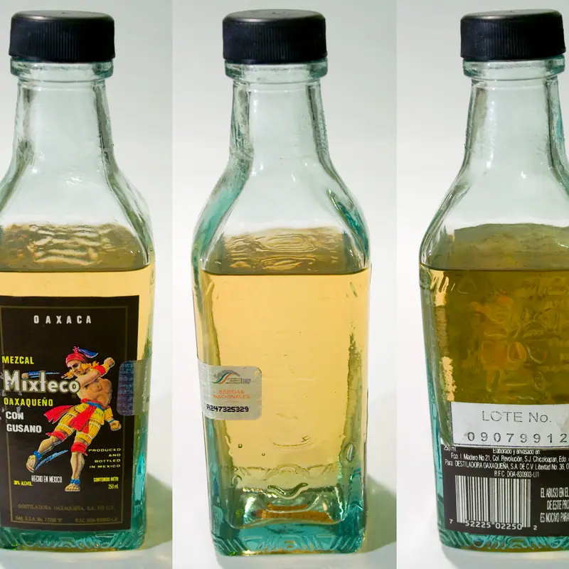 A short glass bottle of pale gold mezcal from Oaxaca, with a black cap and an illustrated label, shown from several angles