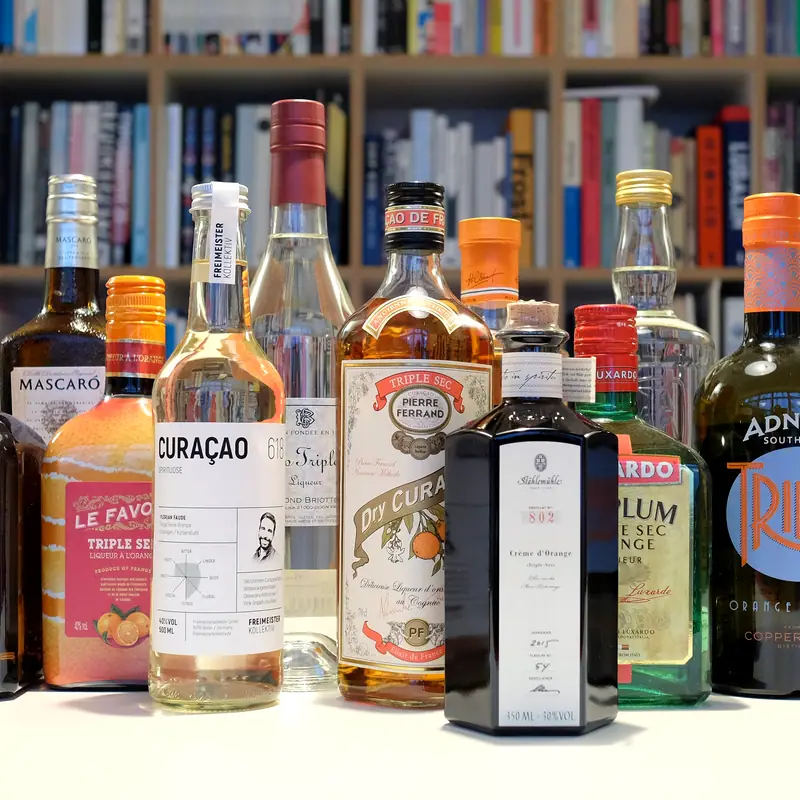 A row of orange curaçao and triple sec bottles, including Pierre Ferrand Dry Curaçao, on a shelf
