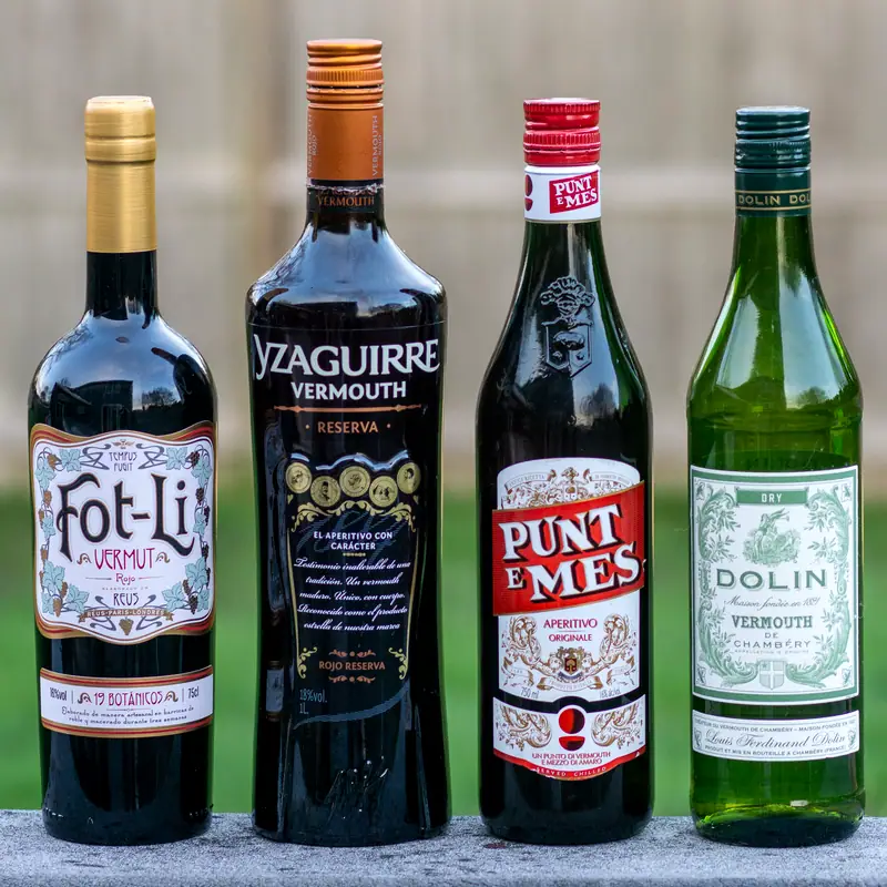 Four bottles of vermouth standing in a row, three dark and one green, each with a different producer's label