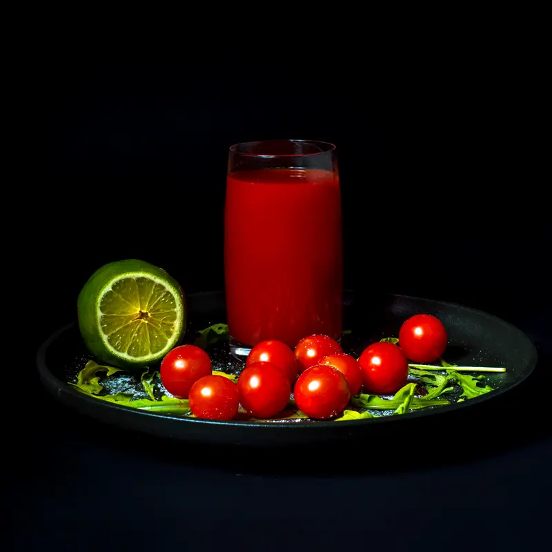 A glass of red tomato juice beside fresh tomatoes and lime on a dark plate