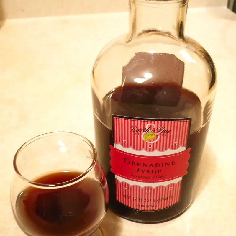 A stoppered glass bottle of dark red grenadine beside a small glass holding a measure of it